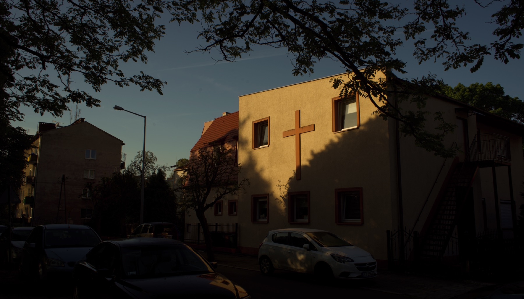Żółty dwupiętrowy budynek z krzyżem na ścianie, oświetlony niskim słońcem. Przed nim ulica z zaparkowanymi samochodami.
